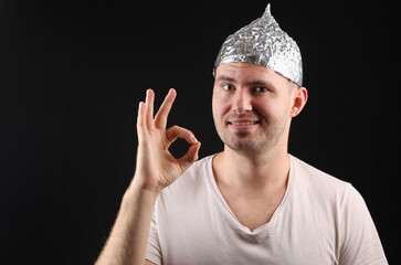 Cheerful Man in foil hat smiles and shows okay on black background