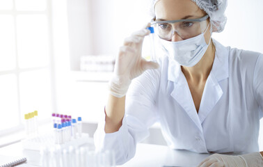 Professional female scientist in protective eyeglasses researching tube with reagents in sunny...