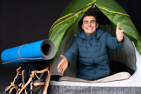 Teenager caucasian man inside a camping green tent isolated on black background presenting and inviting to come with hand