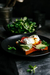 Breakfast. Sandwich with ham, poached egg, avocado and spinach in a plate on a dark background