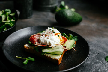 Breakfast. Sandwich with ham, poached egg, avocado and spinach in a plate on a dark background