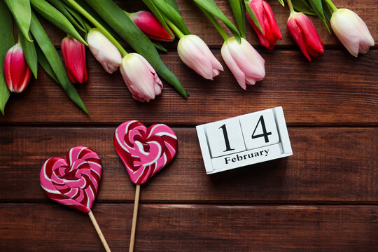 Valentine's Day concept. Wooden calendar 14 February, beautiful tulips flowers and heart shaped candies on dark wooden background. Festive, holiday composition. Top view, flat lay