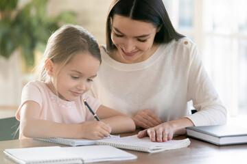 Smiling young mother and little 7s daughter study at home write make homework together. Happy Caucasian mom and small girl child learn distant handwrite prepare school home task or assignment.