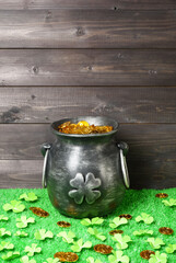 Cast iron pot, decorated by four-petal lucky shamrock leaf, full of leprechaun gold treasure, clover leaves and gold coins on green grass, dark wooden planks background. Saint Patricks Day banner