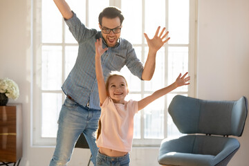 Fototapeta na wymiar Happy young Caucasian father and small 7s daughter have fun dancing listening to music at home. Overjoyed dad and little girl child engaged in funny game together, enjoy family weekend indoors.