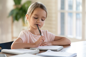 Pensive small cute Caucasian 7s girl sit at desk at home do homework alone write in notebook...