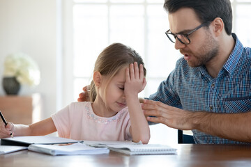 Loving young Caucasian father support comfort unhappy sad little 7s daughter studying doing homework together. Caring dad cheer upset depressed small girl child stressed with school preparation.