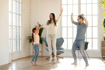 Overjoyed young Caucasian family renters with little 7s daughter have fun dancing together in cozy living room on weekend. Happy mom and dad enjoy family time with small girl child moving to new home.