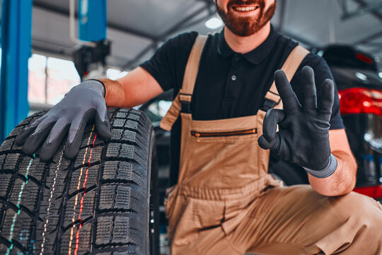 Trustworthy mechanic showing ok gesture, holding car tire and smiling on the car repair shop.