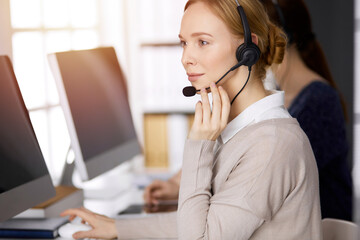 Businesswoman talking by headset while sitting in sunny office. Call center and diverse people group in business