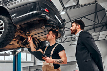 A professional mechanical employee of the car service workshop stand with the male customer under her car on the hydraulic ramp and show her defects and things they have to repair.
