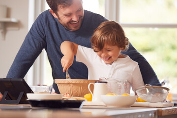 Father And Son In Pyjamas Making Pancakes In Kitchen At Home Following Recipe On Digital Tablet