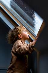 Little preschool boy staying home in bad weather and looking  with interest through window on raindrops and hail, idoors.