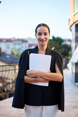 Fototapeta premium Young brunette business woman, wearing stylish black jacket and white dress, holding sheets of paper,standing on balcony. Female portrait in summer. Fashion model on terrace with beautiful city view.