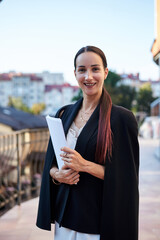 Young brunette business woman, wearing stylish black jacket and white dress, holding sheets of paper,standing on balcony. Female portrait in summer. Fashion model on terrace with beautiful city view.