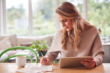 Woman With Digital Tablet Sitting At Table At Home Reviewing Domestic Finances