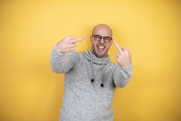 Young bald man wearing glasses over yellow background showing middle finger doing fuck you bad...