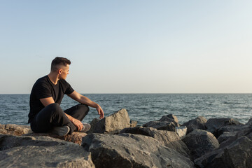 Fototapeta na wymiar Pensive sports man sits on the coast with stones