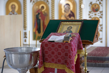 The interior of a Christian church. Prayer table covered with patterned red cloth. A metal container with holy water for baptism. Bible, casket, icon and blurred iconostasis in the background.