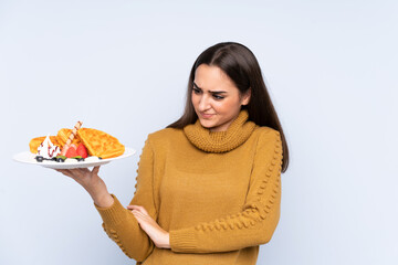 Young caucasian woman holding waffles isolated on blue background with sad expression