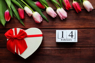 Wooden calendar 14 February, beautiful tulips flowers and heart shaped gift box on dark wooden background. Valentine's Day greeting card. Festive, holiday composition. Top view, flat lay