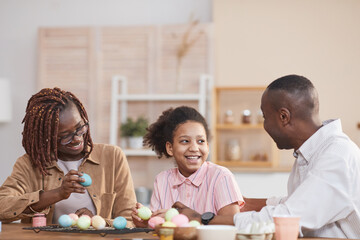Portrait of laughing African-American family painting Easter eggs together while sitting at wooden...