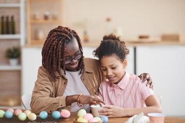 Obraz na płótnie Canvas Portrait of loving African-American mother and daughter painting Easter eggs together while sitting at wooden table in cozy home interior and enjoying DIY art