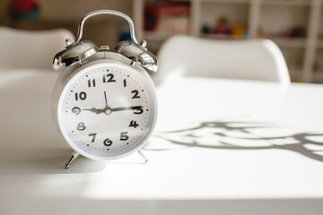 White alarm clock in retro style on a white table in the living room. Seasonal time change, daylight saving time concept.