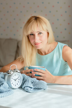 Young sleepy woman holding a cup of coffee. Gray knitted plaid and alarm clock on a white background. Seasonal time change, daylight saving time concept. Vertical image.