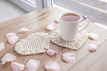 Fototapeta na wymiar Macrame handmade Hobby. Tea in a cup on white macrame coaster on wooden table with rose petals. Food stylist. Eco macrame home decoration. St. Valentine's Day