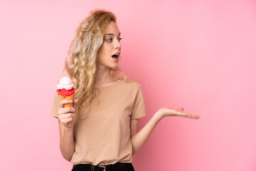 Young blonde woman holding a cornet ice cream isolated on pink background with surprise facial...