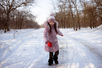 Happy little girl in a down jacket, knitted hat and red mittens runs along the road in a snowy forest. Winter outerwear.