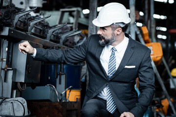 Chief engineer is sitting and checking the quality of the machine at the industry or factory before the engines are starting and running. He is wearing suit and hard hat.