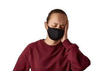 Stress. Young european woman wears face mask and burgundy sweatshirt, suffers from headache and stress, failed a project, tired of work, holds one hand on her head, isolated over white background.