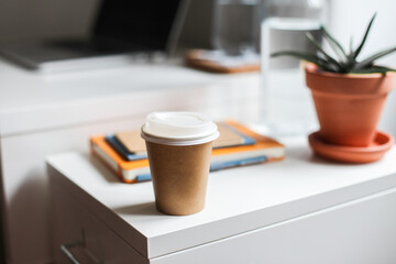 A take away paper coffee cup next to a laptop, notebooks, a glass of water, an aloe plant on a white table