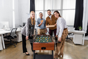 Smiling interracial business people with pizza looking at camera near table soccer in office