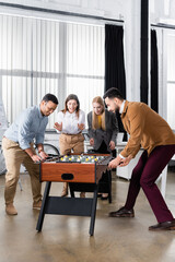 Multiethnic businessmen playing table soccer near businesswomen in office