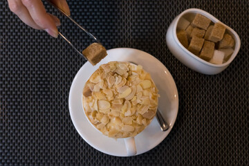 Cup of coffee sprinkled with almonds and hand with tongs with sugar in pieces