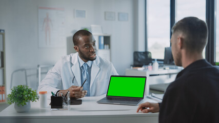 African American Medical Doctor is Explaining Diagnosis to a Patient on a Computer with Green Screen in a Health Clinic. Assistant in White Lab Coat is Reading Medical History in Hospital Office. 