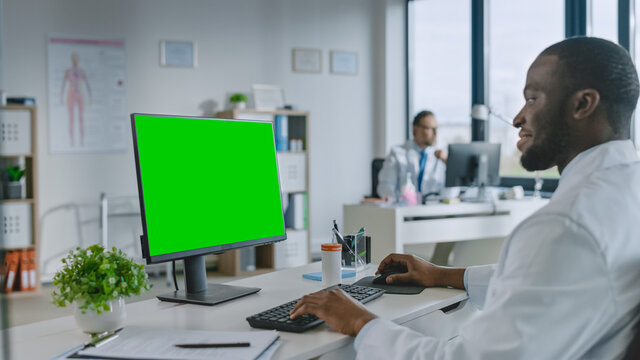 African American Medical Doctor is Working on a Computer with Green Screen Mock Up Display in a Health Clinic. Assistant in White Lab Coat is Reading Medical History Behind a Desk in Hospital Office. - Powered by Adobe