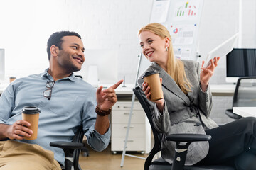 Cheerful indian businessman with coffee to go pointing at colleague with digital tablet