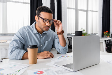 Obraz premium Indian businessman looking at laptop near coffee to go and papers on blurred foreground