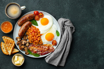 Full English breakfast on a plate with fried eggs, sausages, bacon, beans, toasts and coffee on dark stone background. With copy space. Top view