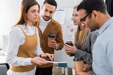 Businesswoman pointing at digital tablet near multicultural colleagues with coffee to go