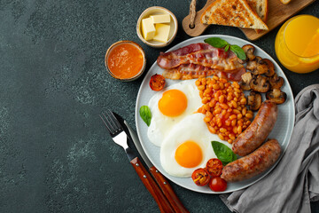 Full English breakfast on a plate with fried eggs, sausages, bacon, beans, toasts and coffee on dark stone background. With copy space. Top view