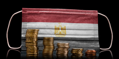 Medical mask with the flag of Egypt behind some shrinking stacks of various coins.(series)
