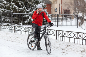 Bicyclist call with smartphone while standing with bike on snowy urban street, riding on bicycle at winter season