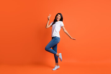 Fototapeta na wymiar Full size photo of optimistic girl stand show v-sign wear white t-shirt jeans sneakers isolated on orange color background