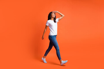 Fototapeta na wymiar Full length photo of girl walk look empty space palm forehead wear white t-shirt jeans sneakers isolated orange color background