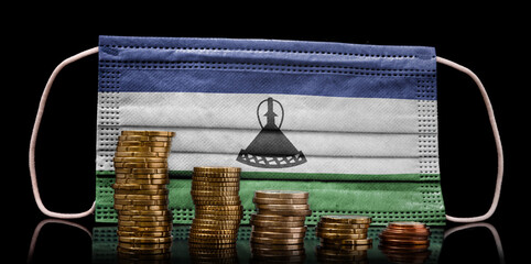 Medical mask with the flag of Lesotho behind some shrinking stacks of various coins.(series)
